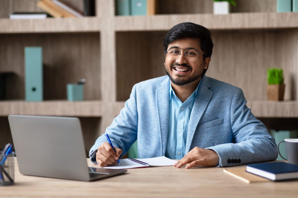 A smiling Indian man with a beard and glasses sits at a desk in a modern office, working on a notebook while looking directly at the camera. He is wearing a blue suit jacket and a light blue shirt.